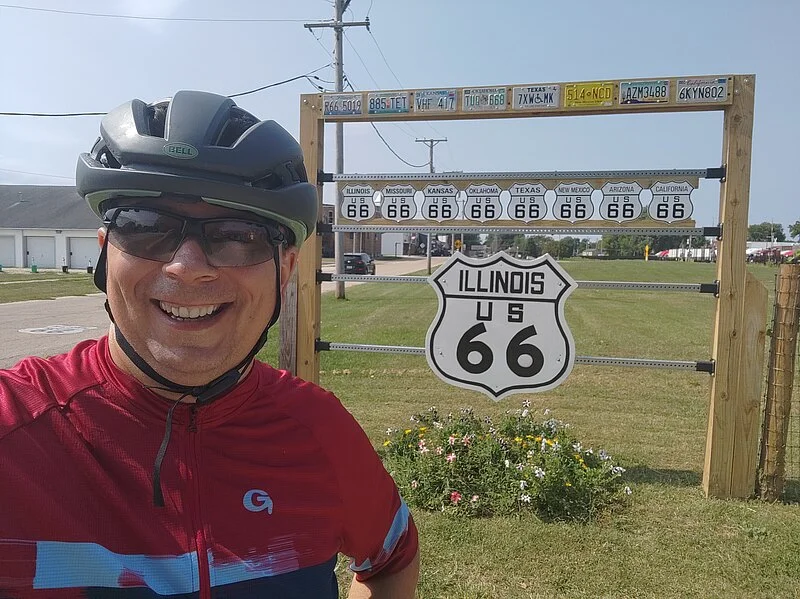 Ein Foto von mir mit Velohelm und Velobrille auf der Route 66. Im Hintergrund befinden sich Route 66 Schilder aller US-Bundesstaaten entlang der Route 66.